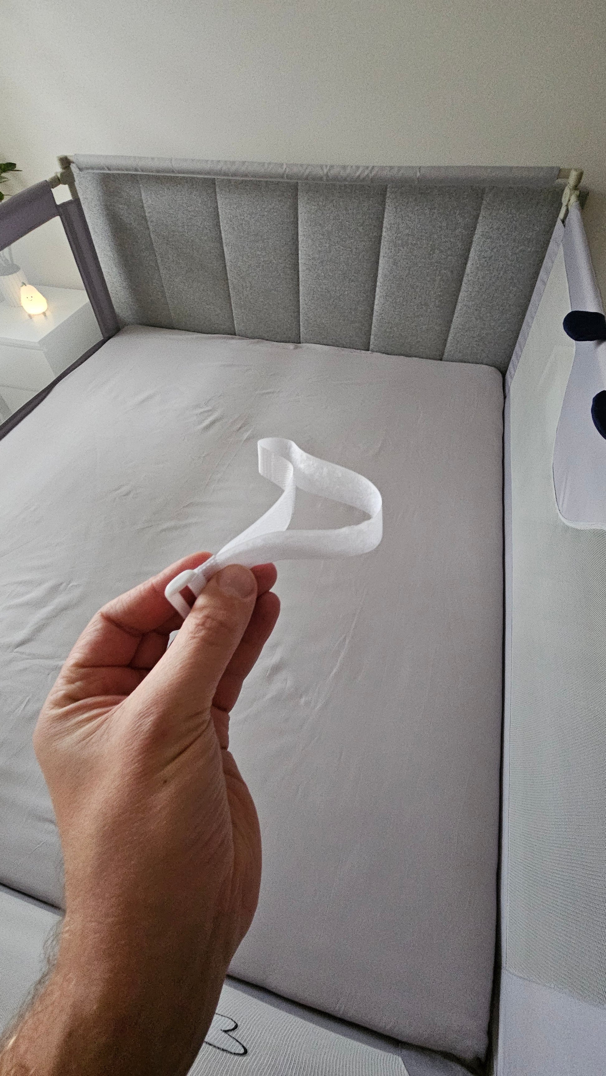 Hand holding white velcro strap above grey baby crib with mesh sides