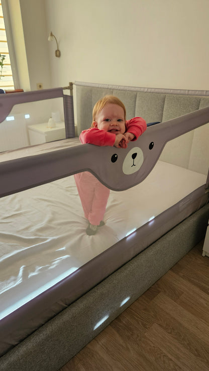 Smiling baby in pink pajamas standing inside gray crib with bear-themed mesh rail in bright nursery