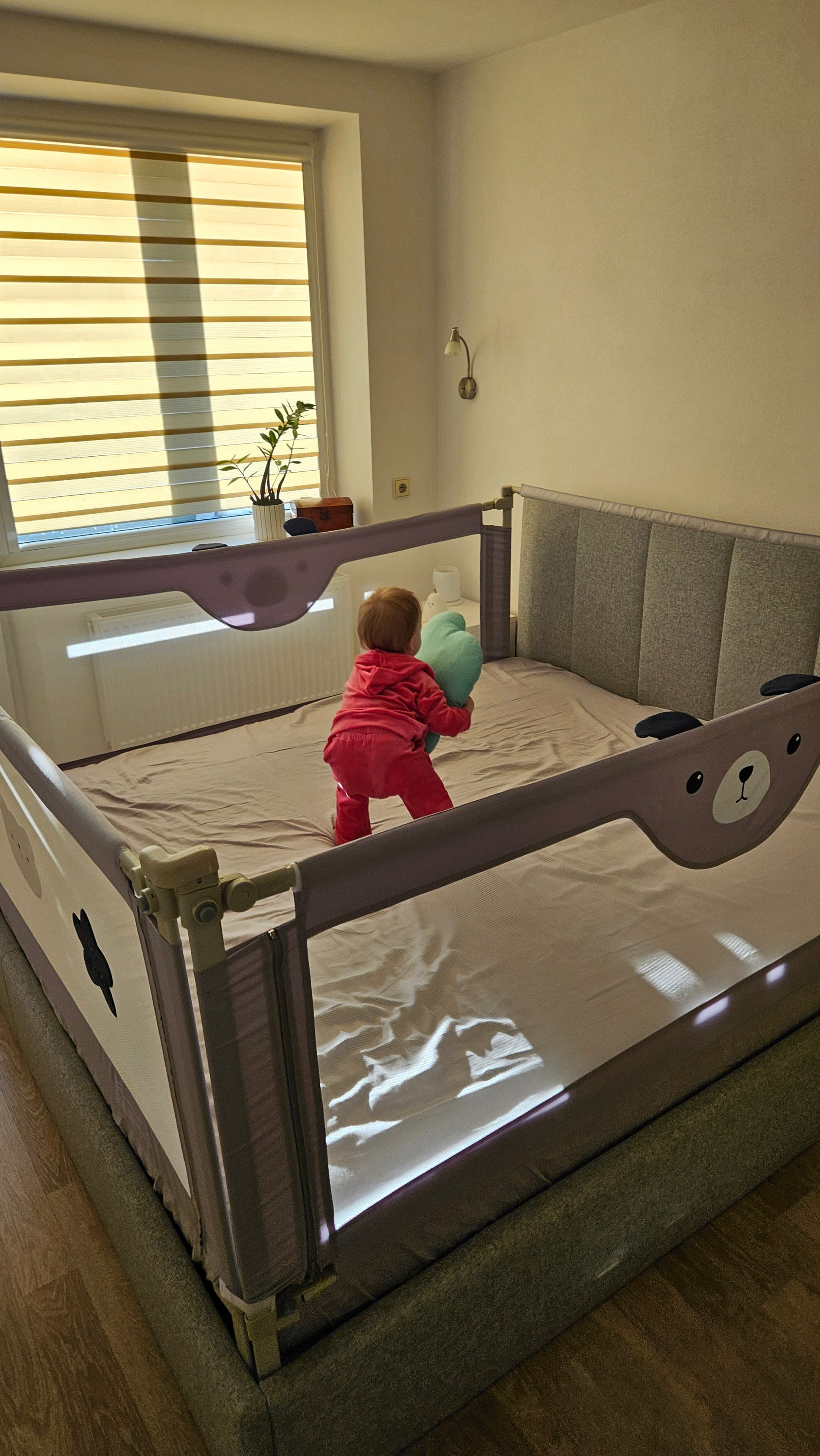 Toddler in pink outfit holding pillow in large bed with safety rails in a sunlit bedroom