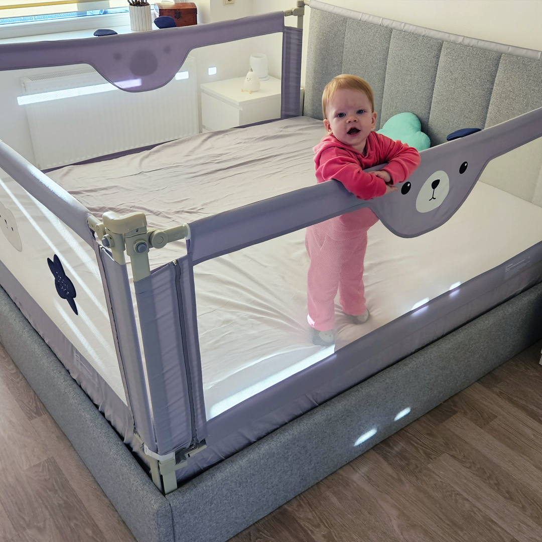 Baby in pink pajamas standing inside a grey bed with child safety rail guard