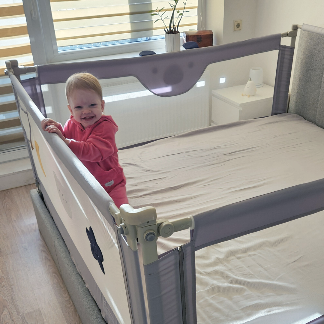 Smiling baby girl in pink onesie standing in a crib with safety bed rail in bright bedroom